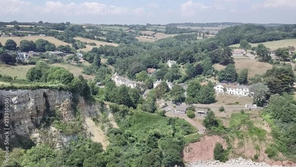 Aerial tilt of a foliage covered cliffside village near the beach on a sunny day. There are several houses in view and open fields on the horizon. Aerial tilt of a foliage covered cliffside village near the beach on a sunny day. There are several houses in view and open fields on the horizon.