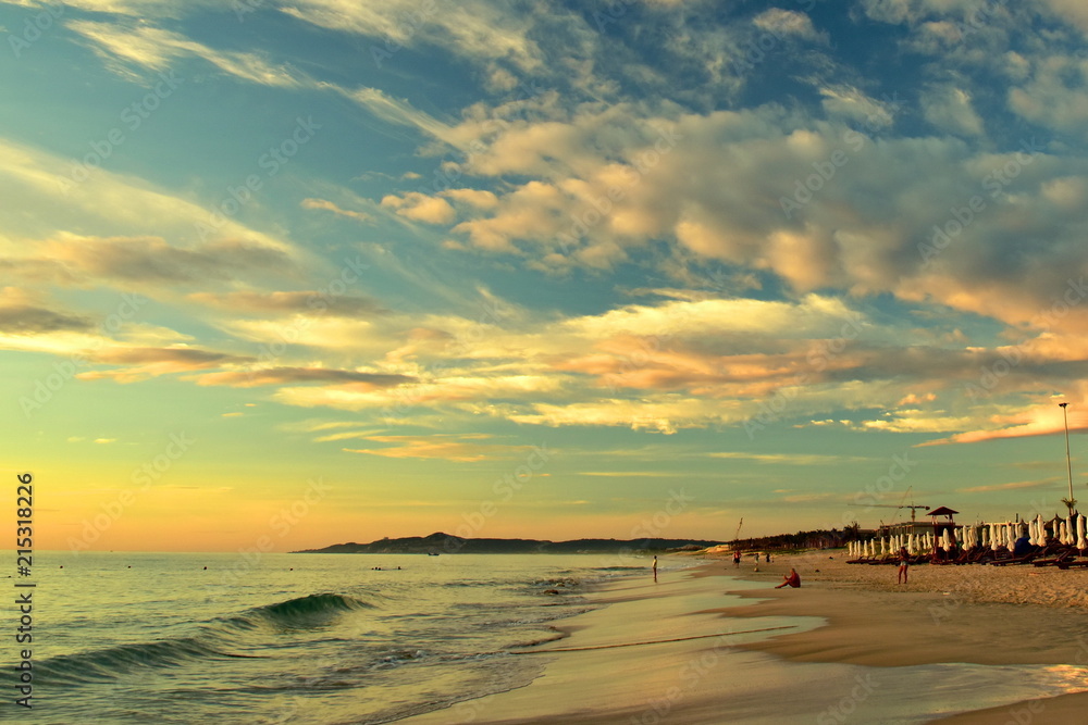 Naklejka premium Sandy beach on the background of a beautiful cloudy sky with golden light. Perspective of the coast. People came to meet the dawn. Beach umbrellas with deckchairs. Waves on the shore