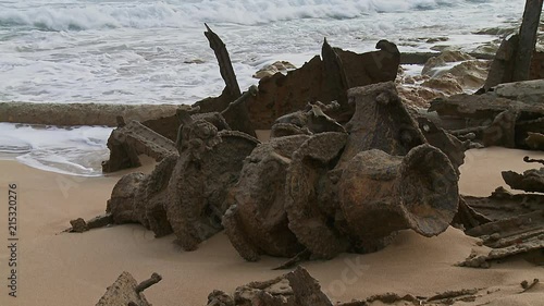 Corroded shaft from a ship wreck on a sandy beach, surrounded by calm waves and other pieces of debris photo