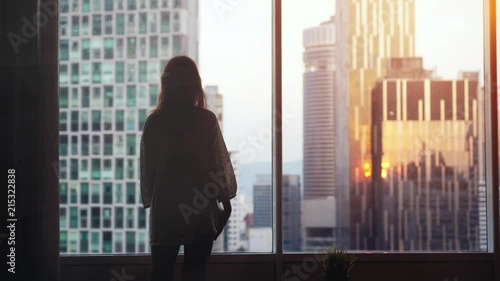 Young pensive woman stands by the big window looking out her apartment on the city buildings during sunset. slow motion. 3840x2160.
