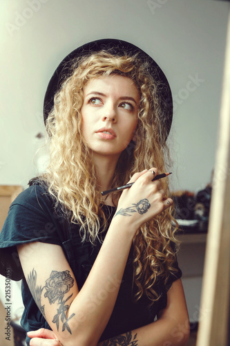 Romantic curly girl in handsome black hat sitting and dreaming with pencil behind the easel in studio
