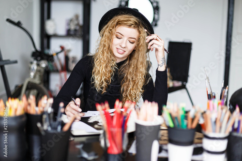 Romantic blonde long hair curly feemale artist in black hat sitting behind the table with color pencil and looking on the sketch in her studo photo