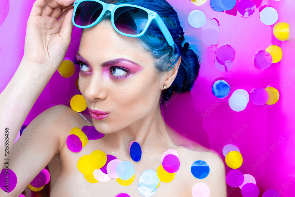 Portrait of young pretty female girl adult woman fashion luxury sexy model with blue hair relaxing wearing turquoise sunglsses in bath isolated by pink water with colorful confetti. Skin care, beauty Portrait of young pretty female girl adult woman fashion luxury sexy model with blue hair relaxing wearing turquoise sunglsses in bath isolated by pink water with colorful confetti. Skin care, beauty