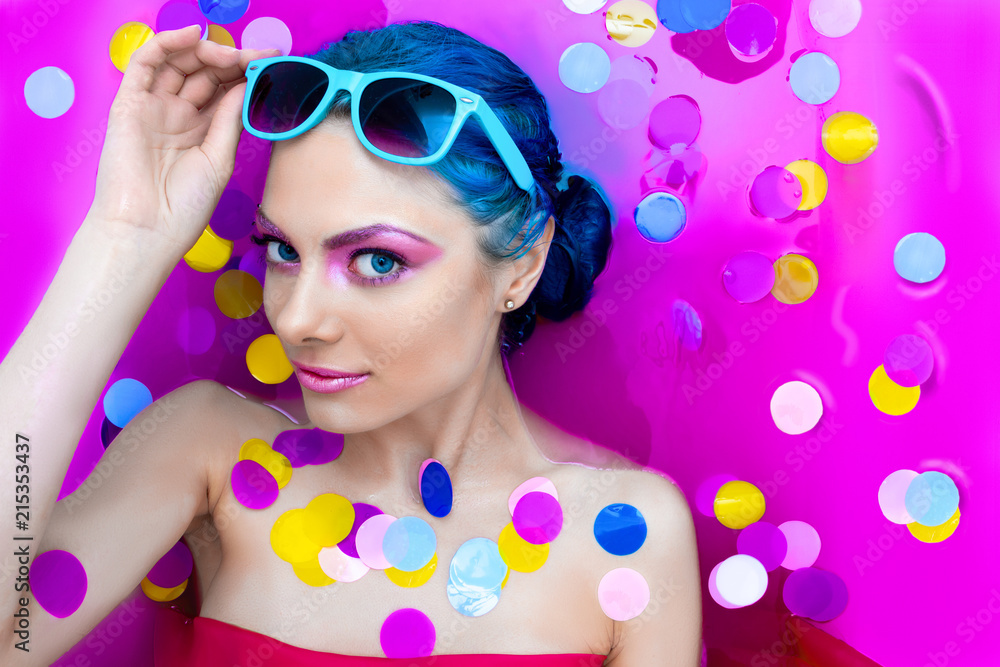 Portrait of young pretty female girl adult woman / fashion luxury sexy model with blue hair relaxing wearing turquoise sunglsses in bath isolated by pink water with colorful confetti. Skin care, beaut Portrait of young pretty female girl adult woman / fashion luxury sexy model with blue hair relaxing wearing turquoise sunglsses in bath isolated by pink water with colorful confetti. Skin care, beaut
