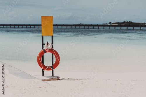 Empty metal mock-up of the warning banner in front of ocean water of a tropical resort with a lifebuoy below; blank orange metallic safety signboard template on the beach with the seascape and bridge