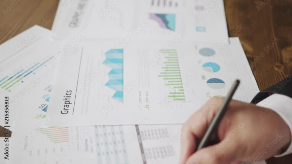 Close up of a businessman hands with pen working at office desk and analyzing graphs and charts and writing a business plan. Trader man monitoring annual business report. Business concept Close up of a businessman hands with pen working at office desk and analyzing graphs and charts and writing a business plan. Trader man monitoring annual business report. Business concept