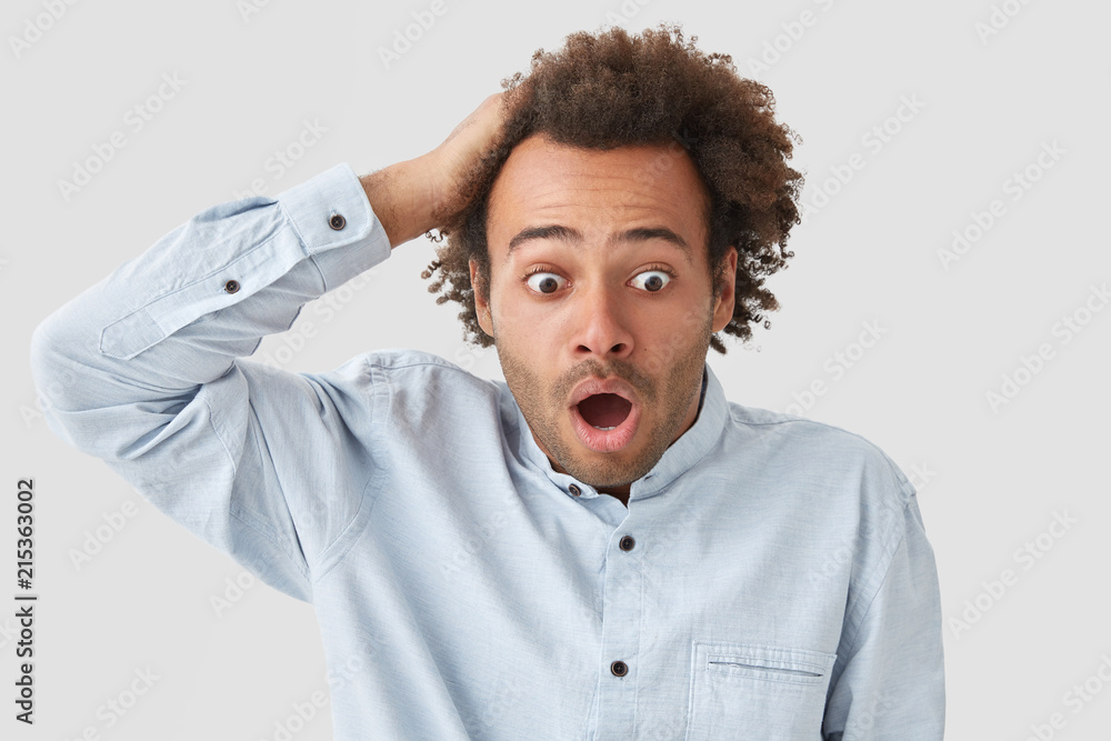Shocked curly male looks with terrified expression, keeps hand on head, stares with bugged eyes, hears something terrible, dressed in white shirt, poses indoor. Puzzled African American guy. Shocked curly male looks with terrified expression, keeps hand on head, stares with bugged eyes, hears something terrible, dressed in white shirt, poses indoor. Puzzled African American guy.