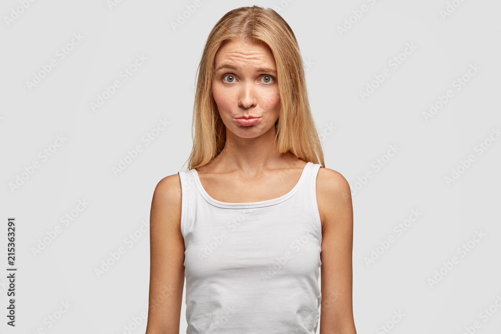 Cute young female purses lower lips and looks with displeased expression, wears casual t shirt, feels offended after quarrel with boyfriend, isolated over white background. People and lifestyle Cute young female purses lower lips and looks with displeased expression, wears casual t shirt, feels offended after quarrel with boyfriend, isolated over white background. People and lifestyle