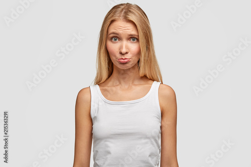 Cute young female purses lower lips and looks with displeased expression, wears casual t shirt, feels offended after quarrel with boyfriend, isolated over white background. People and lifestyle
