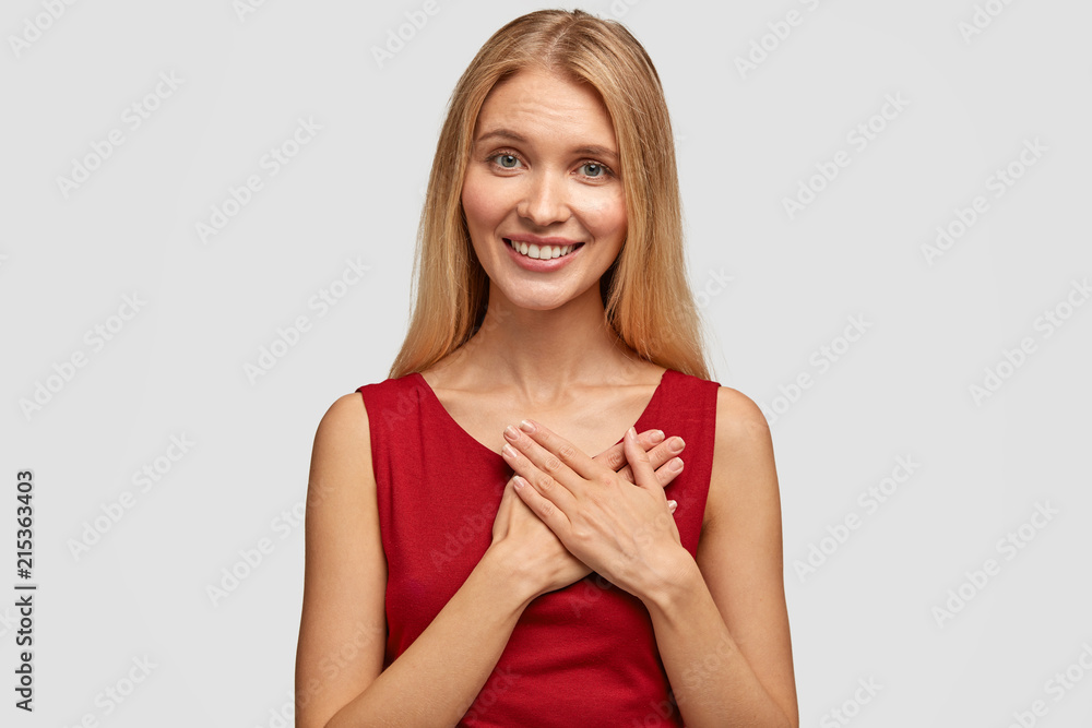 Adorable glad female keeps both palms on heart, expresses good attitude to someone, has appealing look, dressed in casual red vest, isolated over white background, expresses friendly manners Adorable glad female keeps both palms on heart, expresses good attitude to someone, has appealing look, dressed in casual red vest, isolated over white background, expresses friendly manners