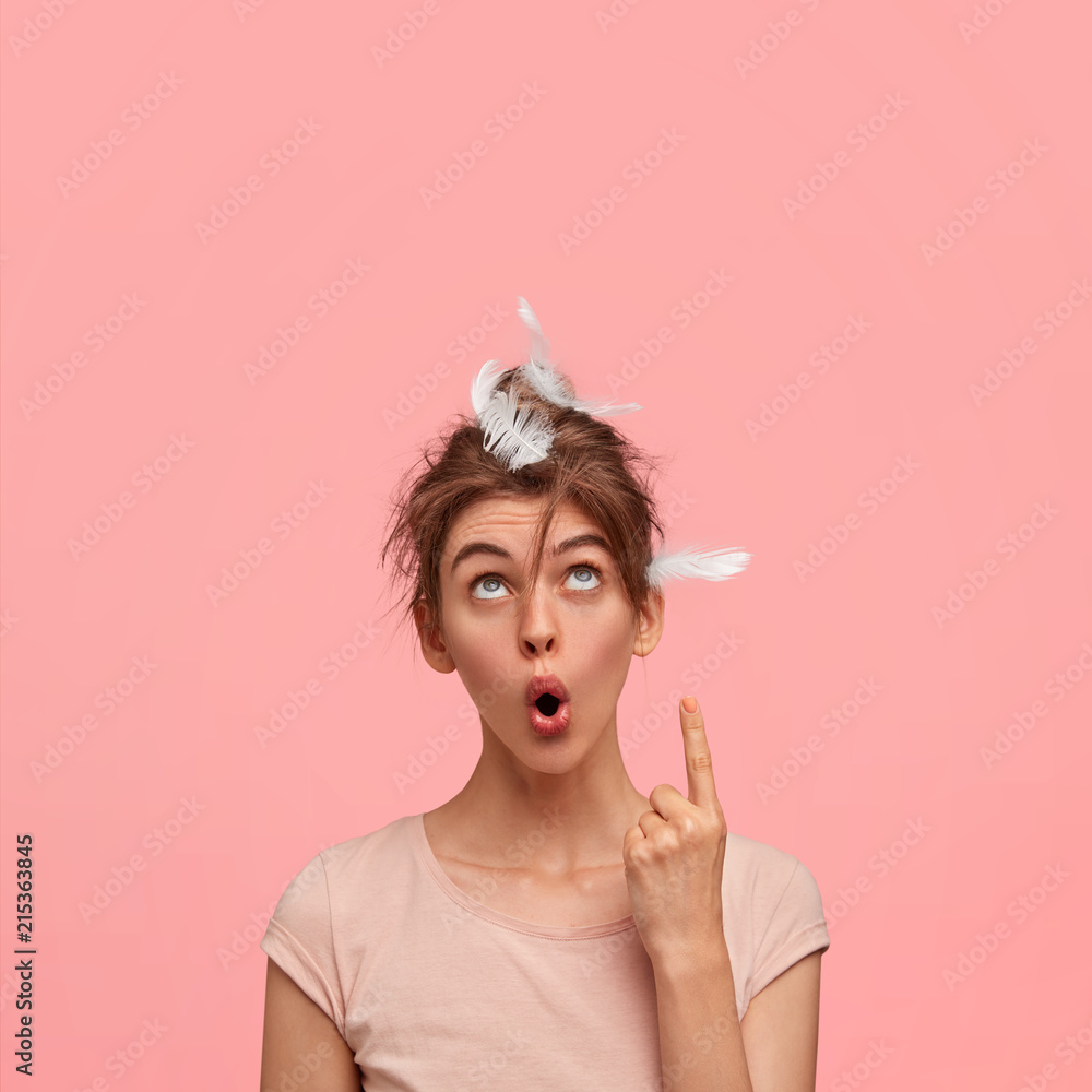 Vertical shot of stupefied sleepy female with feather on head, has astonished facial expression, points upwards, shows free space for your promotional content. Monochrome. Awakening concept. Vertical shot of stupefied sleepy female with feather on head, has astonished facial expression, points upwards, shows free space for your promotional content. Monochrome. Awakening concept.