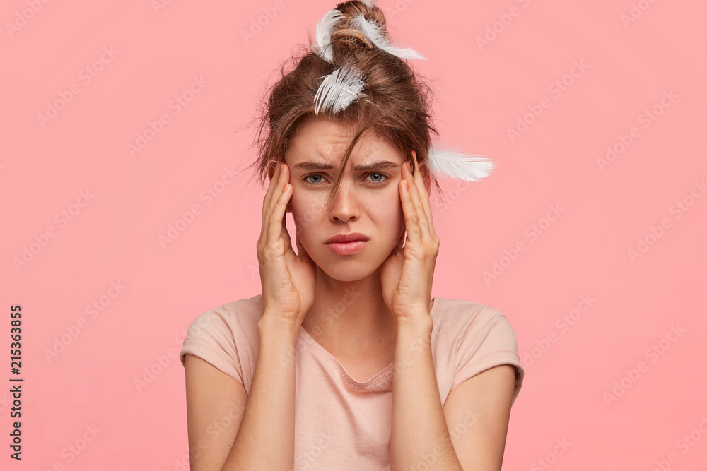 Upset young female keeps hands on temples, suffers from headache, frowns face with displeasure, has bad sleep during night, dressed in casual t shirt, isolated over pink background in bedroom. Upset young female keeps hands on temples, suffers from headache, frowns face with displeasure, has bad sleep during night, dressed in casual t shirt, isolated over pink background in bedroom.