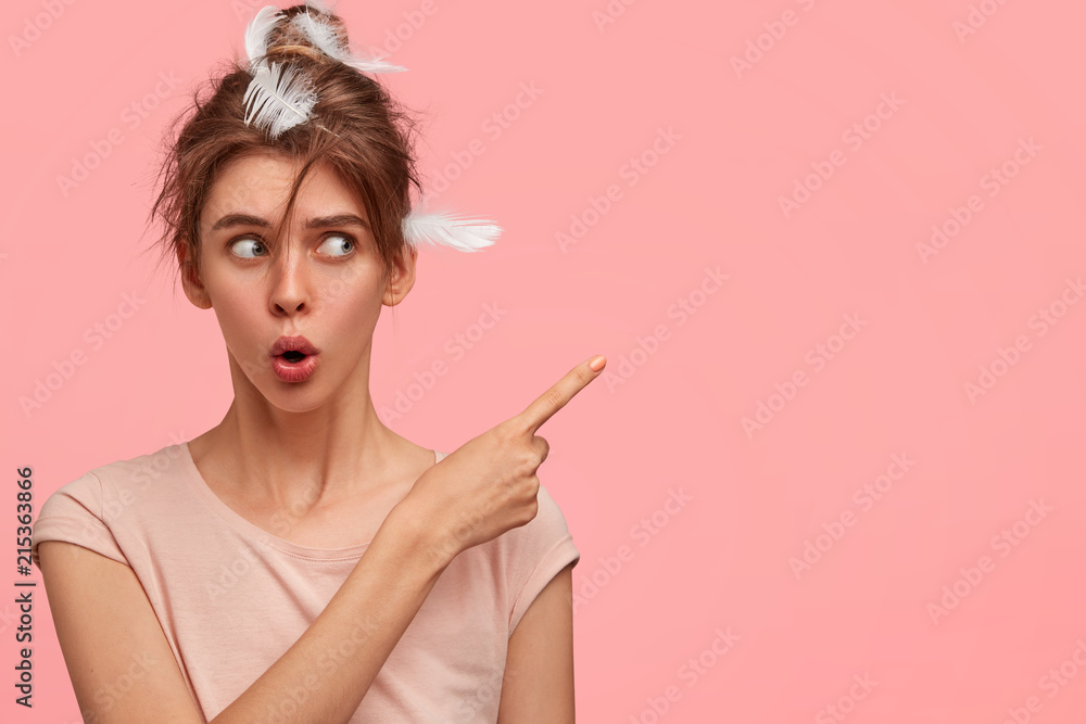 Horizontal shot of shocked female points aside with index finger, has amazed expression, feathers in hair, shows free space for your advertisement or promotional text. People and sleep concept Horizontal shot of shocked female points aside with index finger, has amazed expression, feathers in hair, shows free space for your advertisement or promotional text. People and sleep concept