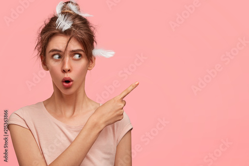 Horizontal shot of shocked female points aside with index finger, has amazed expression, feathers in hair, shows free space for your advertisement or promotional text. People and sleep concept