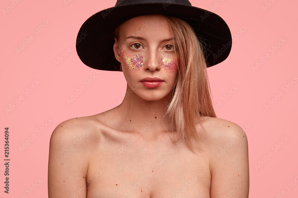 Cropped image of good looking Caucasian female with sparkles on cheeks, has bare shoulders, looks seriously at camera, poses for fashion magazine, isolated over pink background, wears only hat Cropped image of good looking Caucasian female with sparkles on cheeks, has bare shoulders, looks seriously at camera, poses for fashion magazine, isolated over pink background, wears only hat