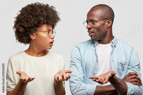 Confused African American couple look at each other angrily, clasp hands, don`t know where their money disappeared, wear round spectacles, isolated over white background. Family has problems
