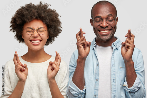Cheerful African American female and male students cross fingers, haave broad smiles, hope for good luck before exam, isolated over white background. People, ethnicity and body language concept photo