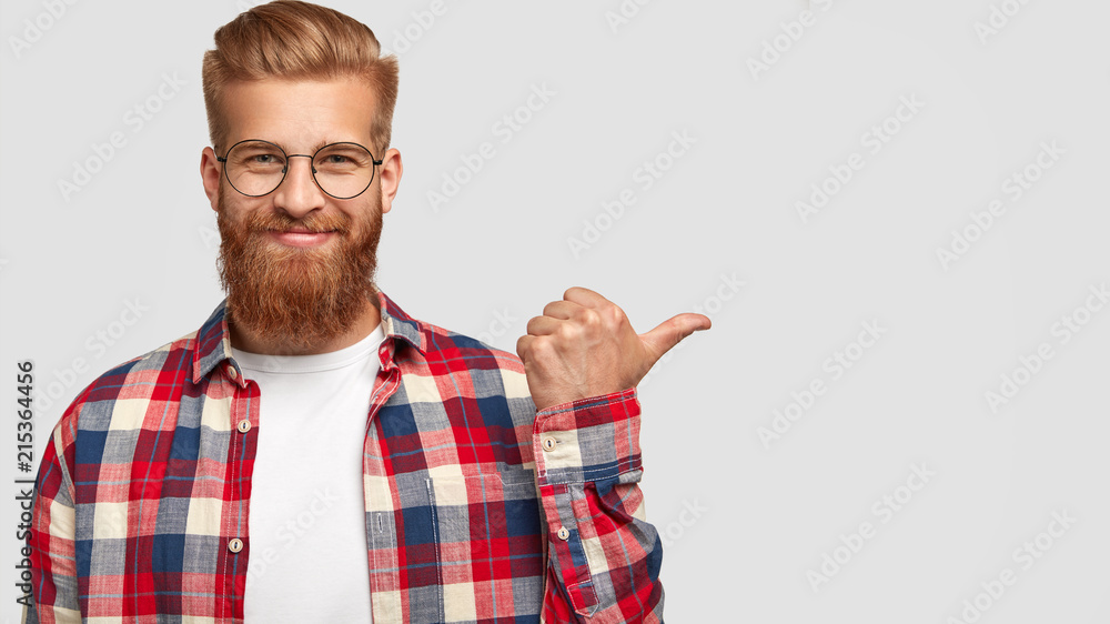 Cheerful bearded male has charming smile, points aside, shows nice place to visit, attracts your attention, dressed in stylish clothes, has trendy hairstyle. People, style, advertisement concept Cheerful bearded male has charming smile, points aside, shows nice place to visit, attracts your attention, dressed in stylish clothes, has trendy hairstyle. People, style, advertisement concept
