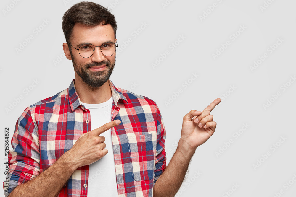 Studio shot of good looking man with satisfied expression, has dark stubble, indicates with fore fingers aside, shows you interesting place, dressed in bright checkered shirt. Advertisement concept Studio shot of good looking man with satisfied expression, has dark stubble, indicates with fore fingers aside, shows you interesting place, dressed in bright checkered shirt. Advertisement concept