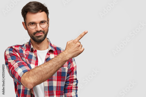 Friendly looking male seller with bristle, dressed in fashionable clothes, points at upper right corner, shows white blank space for your promotional content. People and advertisement concept photo
