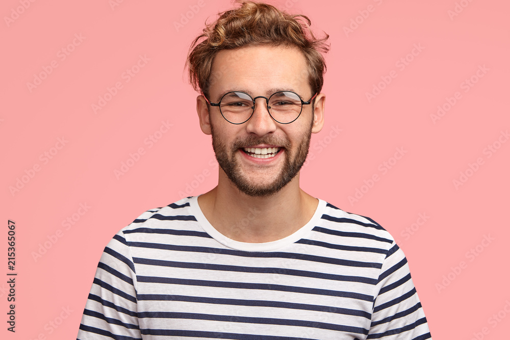 Positive young Caucasian male with pleasant friendly smile, shows white teeth, rejoices new stage in life, wears casual striped sweater and round spectacles, stands alone against pink background. Positive young Caucasian male with pleasant friendly smile, shows white teeth, rejoices new stage in life, wears casual striped sweater and round spectacles, stands alone against pink background.