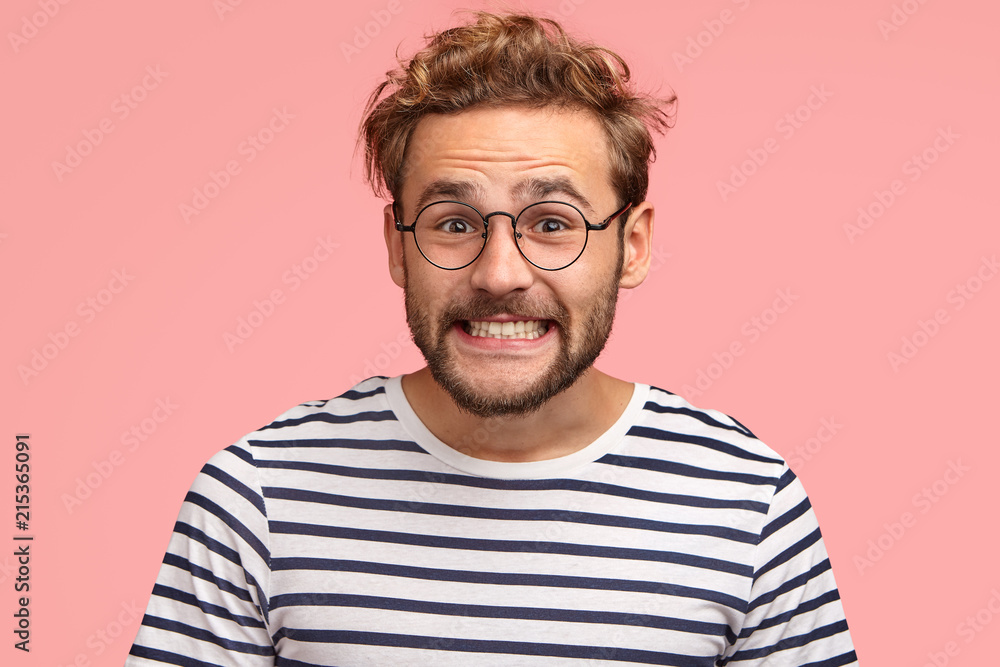 Joyful Caucasian male clenches teeth and looks positively at camera, has curly hair, wears spectacles and striped sweater, isolated over pink background. Happy man freelancer rejoices success - obrazy, fototapety, plakaty 