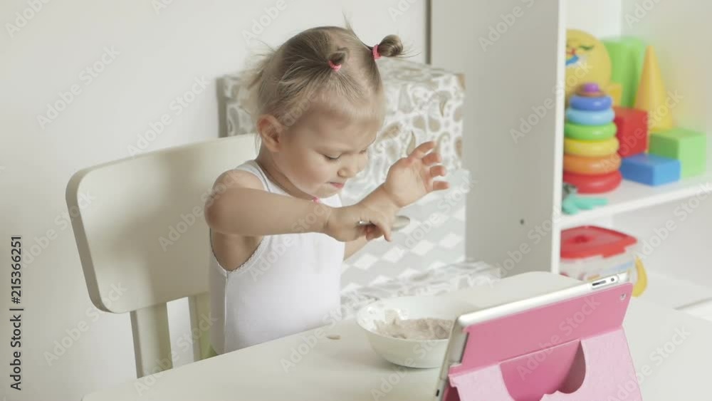 A little girl sitting at a children's table eats porridge with a spoon and looks at the screen of an electronic tablet A little girl sitting at a children's table eats porridge with a spoon and looks at the screen of an electronic tablet