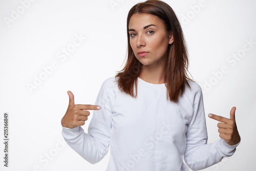 Attractive young female with red hair, points at blank long sleeved t shirt, shows space for advertising content, isolated over white background. People, clothing, fashion, design, promotion concept.