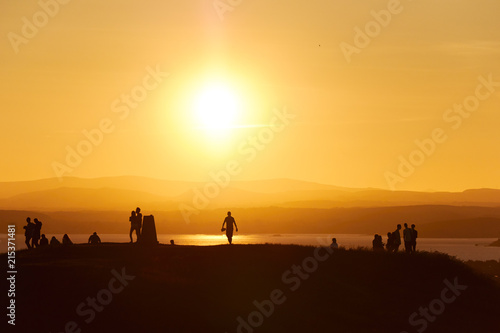 Human silhouettes standing on a hill in front of an orange sunset.