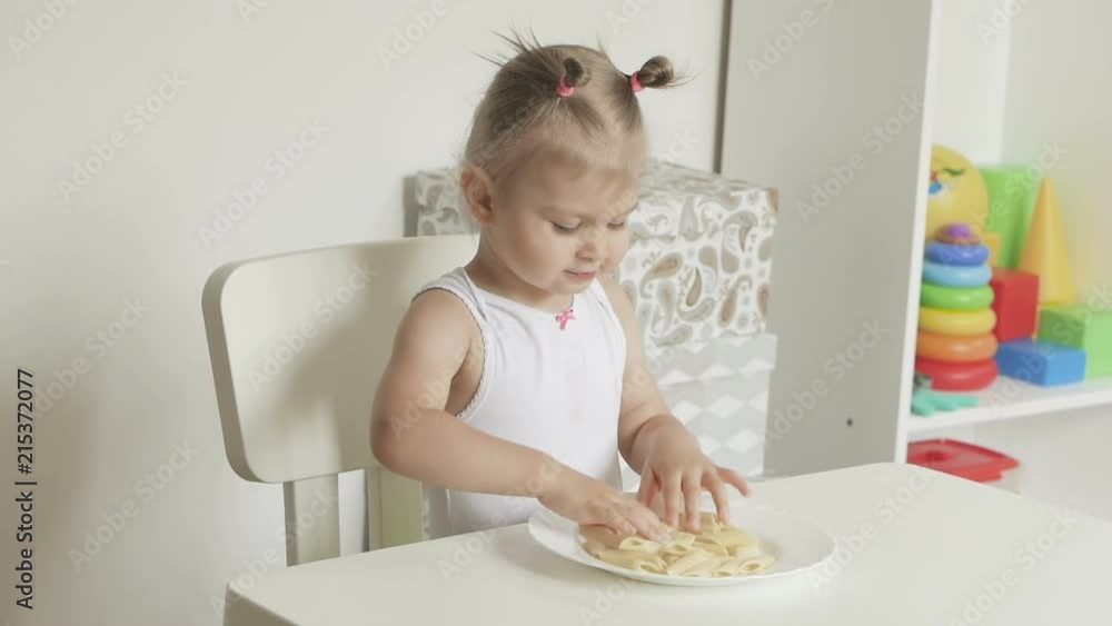 A little girl sitting at a children's table takes her hands and eats pasta A little girl sitting at a children's table takes her hands and eats pasta