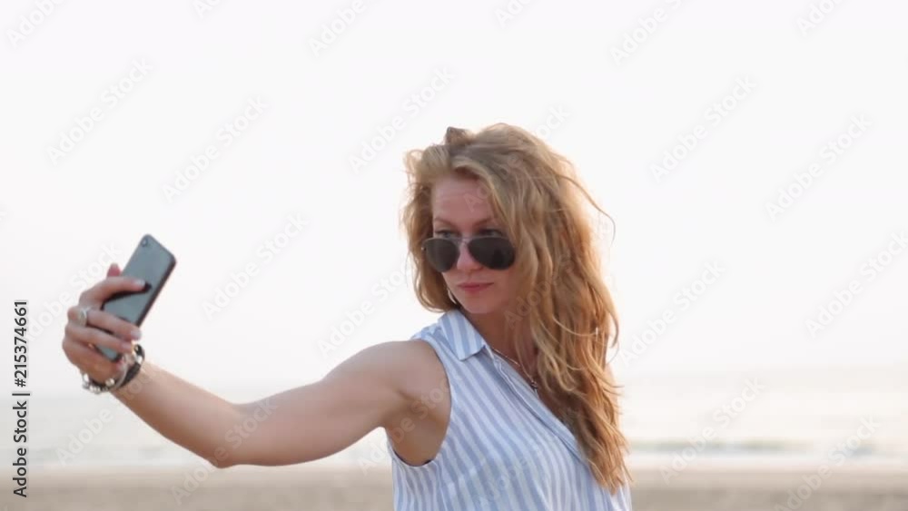 A young sexy blonde girl in sunglasses takes pictures of herself by the phone as a selfie on a sandy beach on the ocean. A young sexy blonde girl in sunglasses takes pictures of herself by the phone as a selfie on a sandy beach on the ocean.