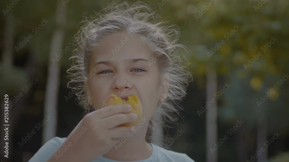 Smiling cute preteen girl eating delicious peach outdoors. Adorable elementary age child enjoying taste of peach and looking at camera with toothy smile during family picnic in park on sunny day. Stock ビデオ | Adobe Stock 