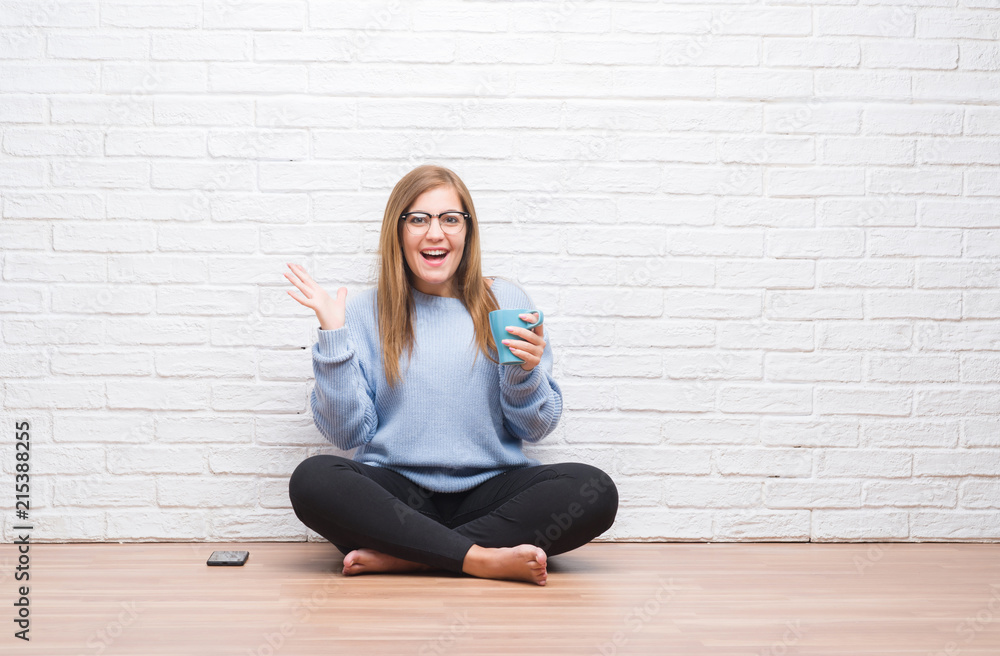 Young adult woman sitting on the floor over white brick wall driking cup of coffee very happy and excited, winner expression celebrating victory screaming with big smile and raised hands Young adult woman sitting on the floor over white brick wall driking cup of coffee very happy and excited, winner expression celebrating victory screaming with big smile and raised hands