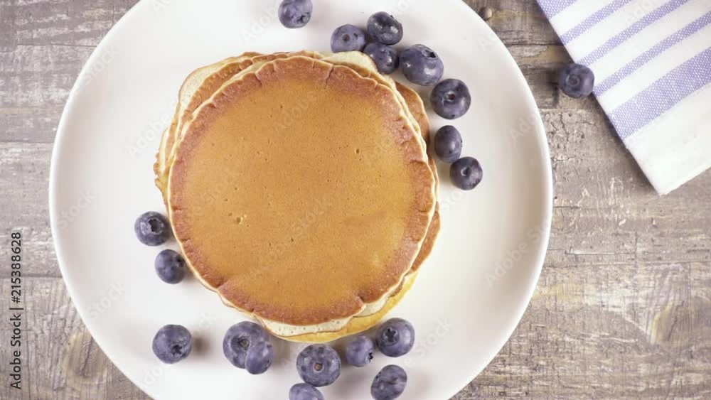 Slow motion blueberries falls on a stack of pancakes on a white plate top view Slow motion blueberries falls on a stack of pancakes on a white plate top view