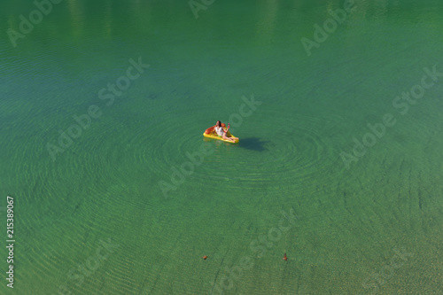 A young woman spends leisure time on an inflatable  sun bed in the middle of a green lake. She wears a white swim suit and enjoys the summer sun as she relaxes on the sun bed. 