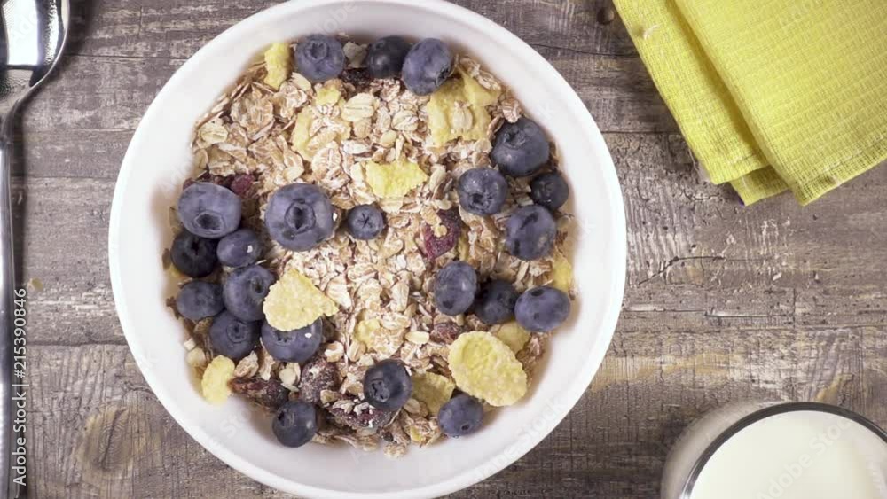 Slow motion fresh blueberries fall into a white bowl of oat and corn flakes with dried fruits top view Slow motion fresh blueberries fall into a white bowl of oat and corn flakes with dried fruits top view
