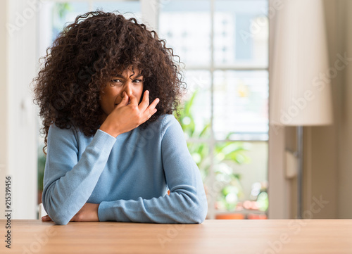 African american woman at home smelling something stinky and disgusting, intolerable smell, holding breath with fingers on nose. Bad smells concept.