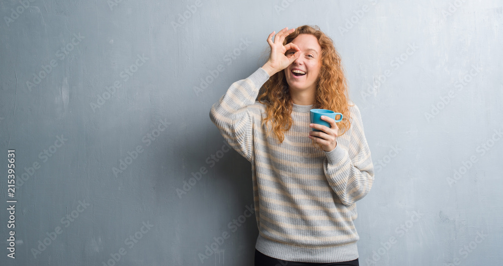 Young redhead woman over grey grunge wall drinking a cup of coffee with happy face smiling doing ok sign with hand on eye looking through fingers Young redhead woman over grey grunge wall drinking a cup of coffee with happy face smiling doing ok sign with hand on eye looking through fingers