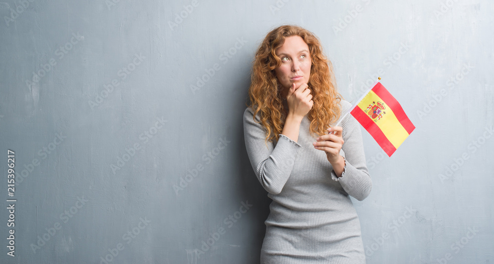 Young redhead woman over grey grunge wall holding flag of Spain serious face thinking about question, very confused idea Young redhead woman over grey grunge wall holding flag of Spain serious face thinking about question, very confused idea