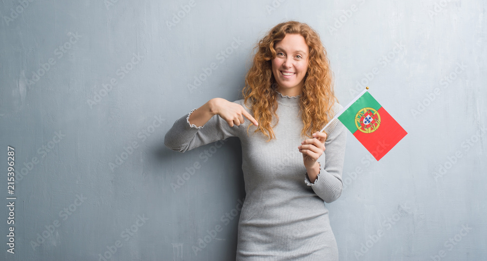 Young redhead woman over grey grunge wall holding flag of Portugal with surprise face pointing finger to himself Young redhead woman over grey grunge wall holding flag of Portugal with surprise face pointing finger to himself