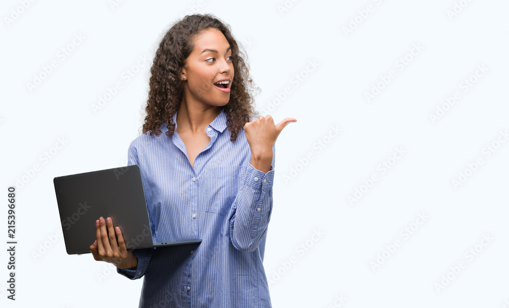 Young hispanic woman holding computer laptop pointing and showing with thumb up to the side with happy face smiling Young hispanic woman holding computer laptop pointing and showing with thumb up to the side with happy face smiling