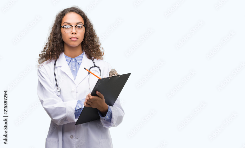 Young hispanic doctor woman holding a clipboard with a confident expression on smart face thinking serious Young hispanic doctor woman holding a clipboard with a confident expression on smart face thinking serious