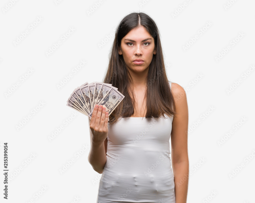 Young hispanic woman holding dollars with a confident expression on smart face thinking serious Young hispanic woman holding dollars with a confident expression on smart face thinking serious