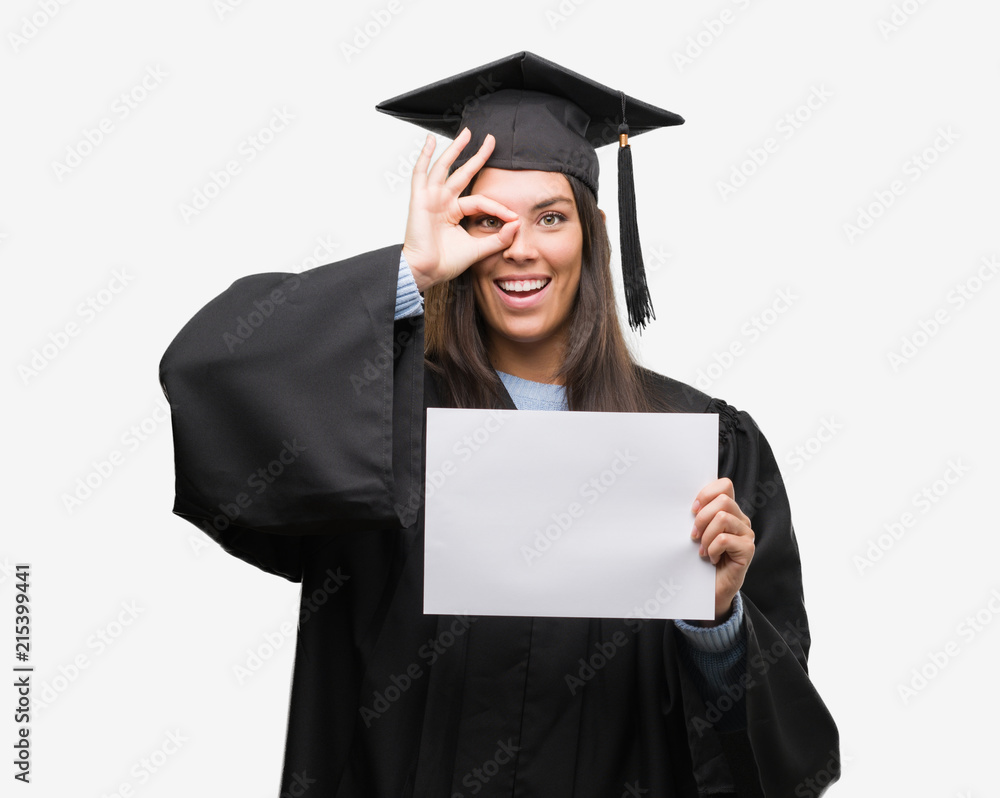 Young hispanic woman wearing graduated uniform holding diploma paper with happy face smiling doing ok sign with hand on eye looking through fingers Young hispanic woman wearing graduated uniform holding diploma paper with happy face smiling doing ok sign with hand on eye looking through fingers