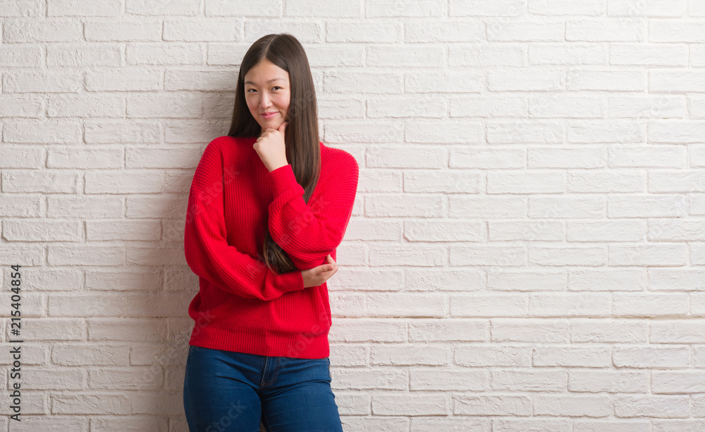 Young Chinese woman over brick wall looking confident at the camera with smile with crossed arms and hand raised on chin. Thinking positive. Young Chinese woman over brick wall looking confident at the camera with smile with crossed arms and hand raised on chin. Thinking positive.