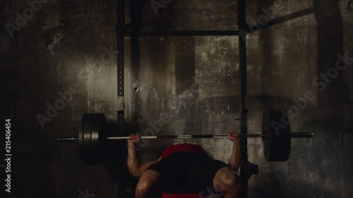 Strong bodybuilder is rising a big rod in a fitness room. He is lying on a training apparatus, putting it off from rack and working out
