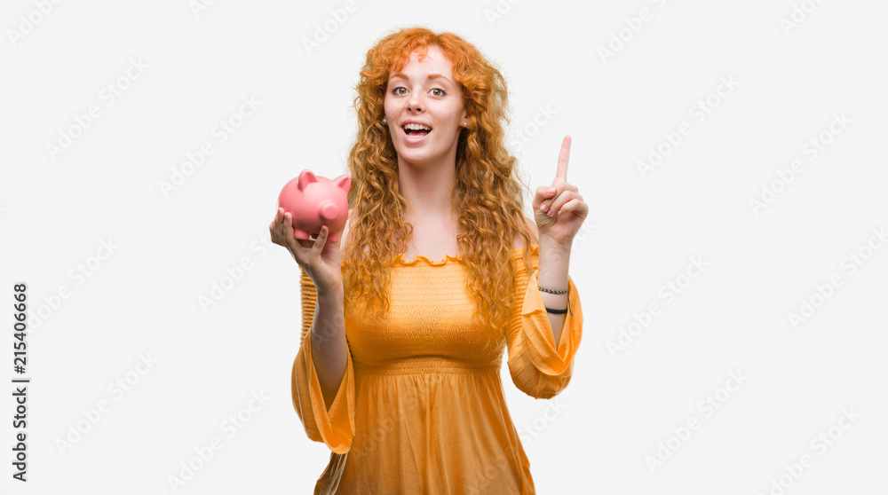 Young redhead woman holding piggy bank surprised with an idea or question pointing finger with happy face, number one