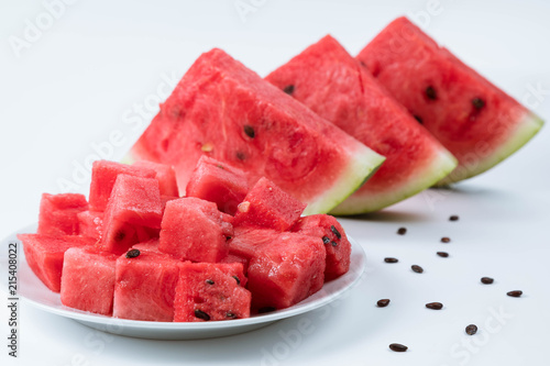 Three segments of watermelon stand on a white table, in the foreground watermelon is cut into cubes. Cubes in soft focus. White background, horizontal shot