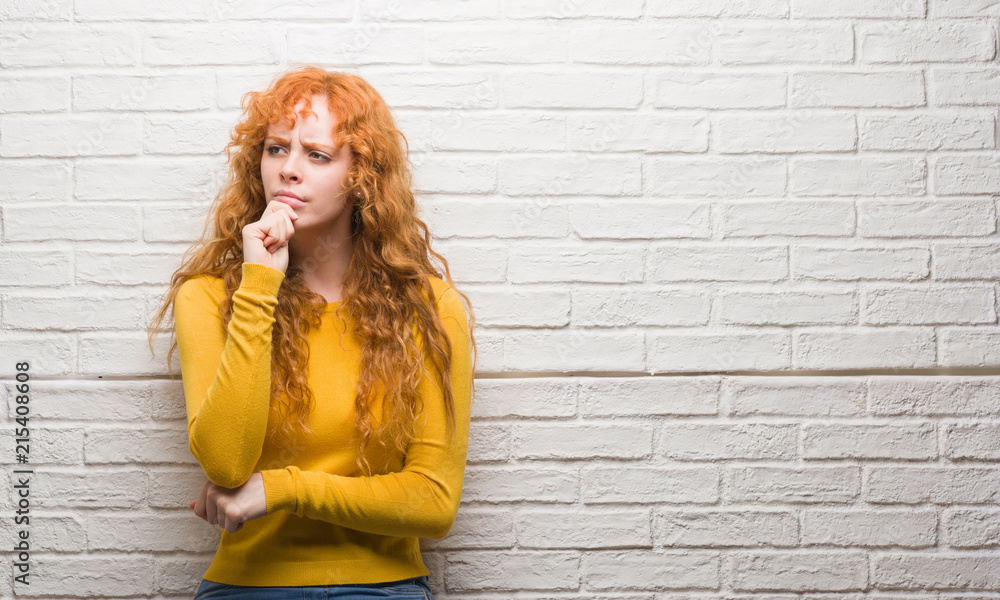 Young redhead woman standing over brick wall serious face thinking about question, very confused idea Young redhead woman standing over brick wall serious face thinking about question, very confused idea
