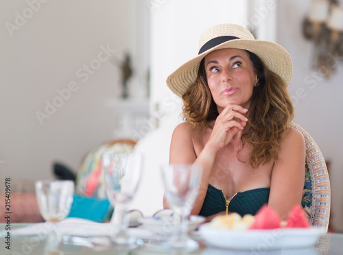 Middle age brunette woman eating wearing summer hat serious face thinking about question, very confused idea
